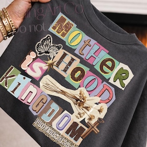 Könnte beinhalten: Dunkelgraues T-Shirt mit dem Schriftzug "Motherhood is Kingdom" im Collage-Stil. Das Design umfasst einen Schmetterling, einen Stern, Blumen und ein Kreuz. Der Text "Proverbs 31:25-27" ist ebenfalls sichtbar.