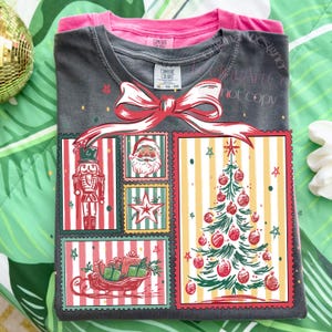 May include: A folded dark gray t-shirt with a Christmas-themed graphic. The graphic features a nutcracker, Santa Claus, a star, a Christmas tree, and a sleigh filled with gifts. A red bow is at the top of the graphic. A pink t-shirt is folded underneath.