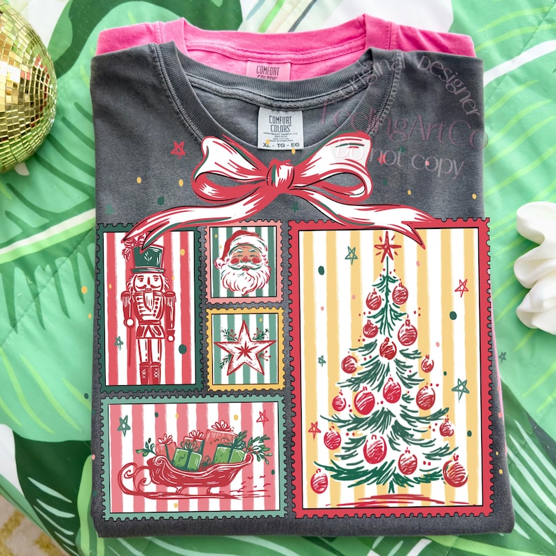 May include: A folded dark gray t-shirt with a Christmas-themed graphic. The graphic features a nutcracker, Santa Claus, a star, a Christmas tree, and a sleigh filled with gifts. A red bow is at the top of the graphic. A pink t-shirt is folded underneath.