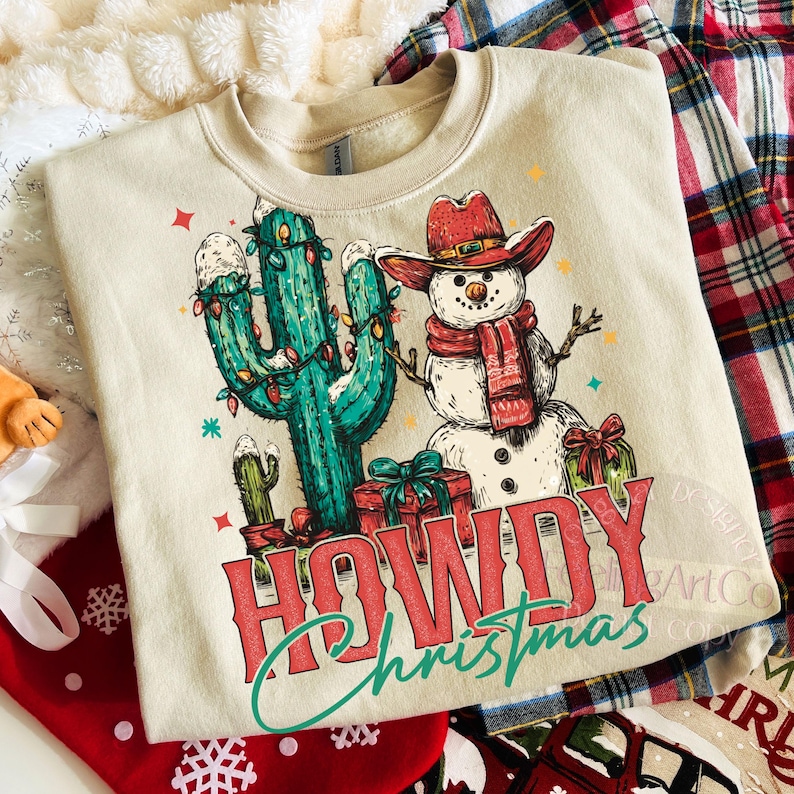Peut inclure: Un sweat-shirt beige avec un motif festif. Le motif repr&eacute;sente un bonhomme de neige portant un chapeau de cowboy et une &eacute;charpe &agrave; c&ocirc;t&eacute; d'un cactus d&eacute;cor&eacute; de lumi&egrave;res de No&euml;l. Les mots "HOWDY Christmas" sont imprim&eacute;s en rouge et vert.