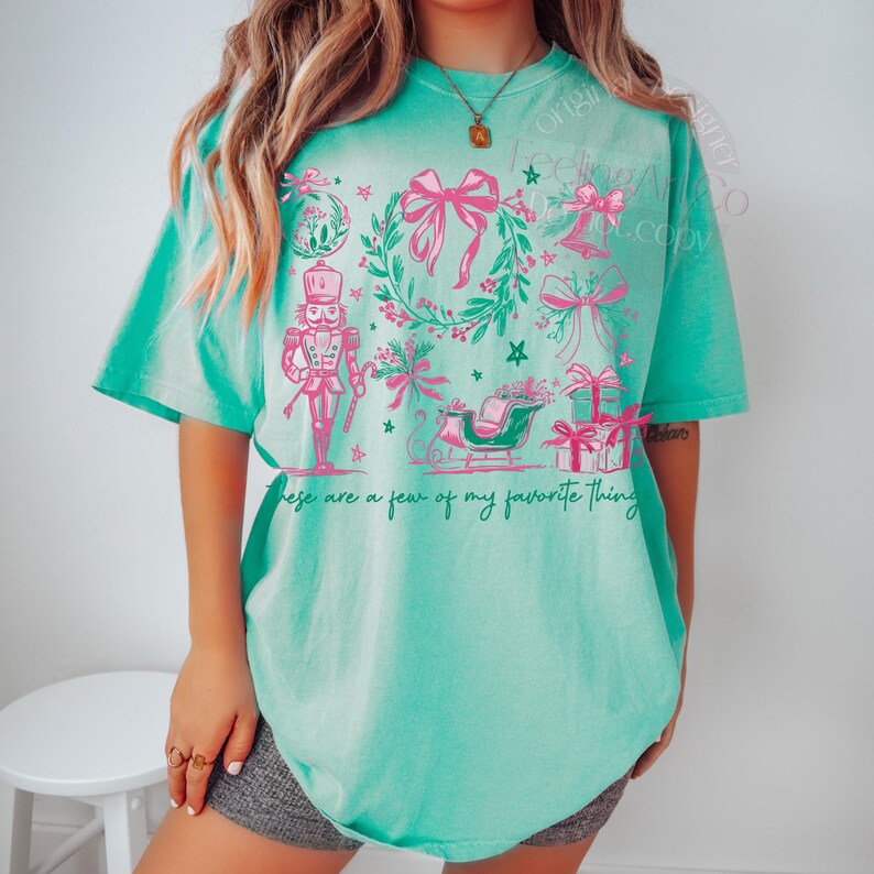 May include: A turquoise t-shirt with a festive design featuring pink bows, a wreath, a nutcracker, a sleigh, and wrapped gifts. The text "These are a few of my favorite things" is printed on the shirt.