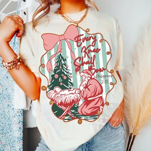 Puede incluir: Camiseta color crema con un dise&ntilde;o navide&ntilde;o. El dise&ntilde;o incluye un lazo rosa, un &aacute;rbol de Navidad y una figura de Pap&aacute; Noel arrodillado. El texto "Every Knee Shall Bow" se muestra dentro de un marco decorativo.