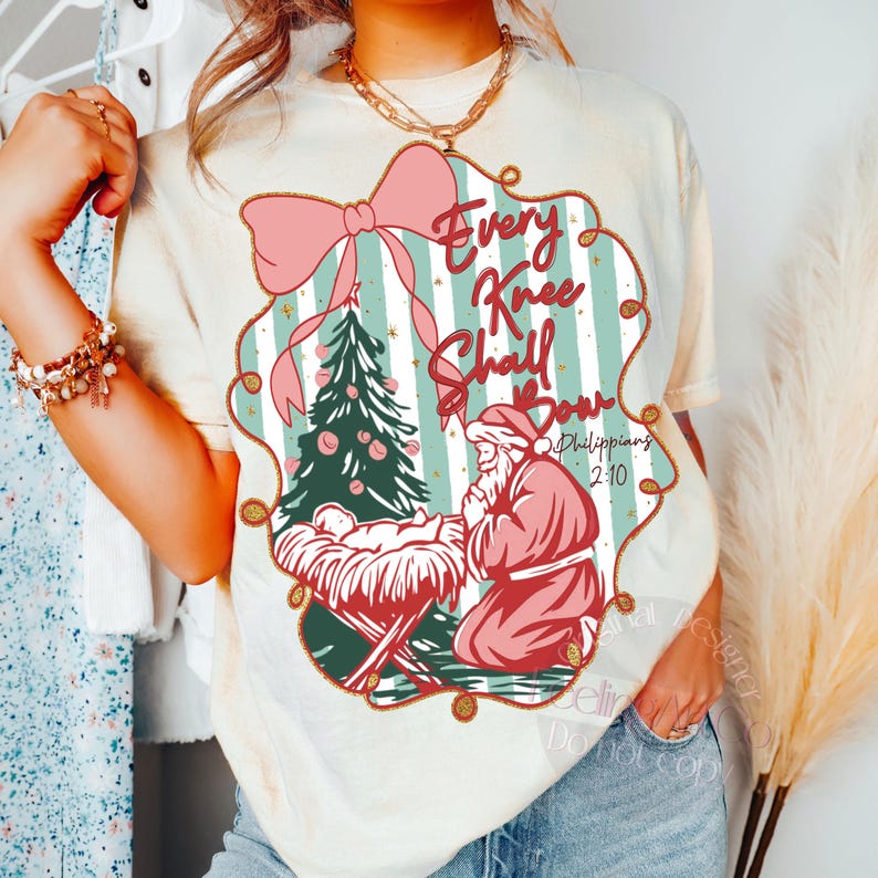 Puede incluir: Camiseta color crema con un dise&ntilde;o navide&ntilde;o. El dise&ntilde;o incluye un lazo rosa, un &aacute;rbol de Navidad y una figura de Pap&aacute; Noel arrodillado. El texto "Every Knee Shall Bow" se muestra dentro de un marco decorativo.