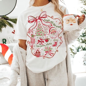 May include: White t-shirt with a festive Christmas design. The graphic includes a red bow, Christmas tree, Santa Claus, and the text "Jingle all the way, Christmas, Please." A Santa mug is held in the hand.