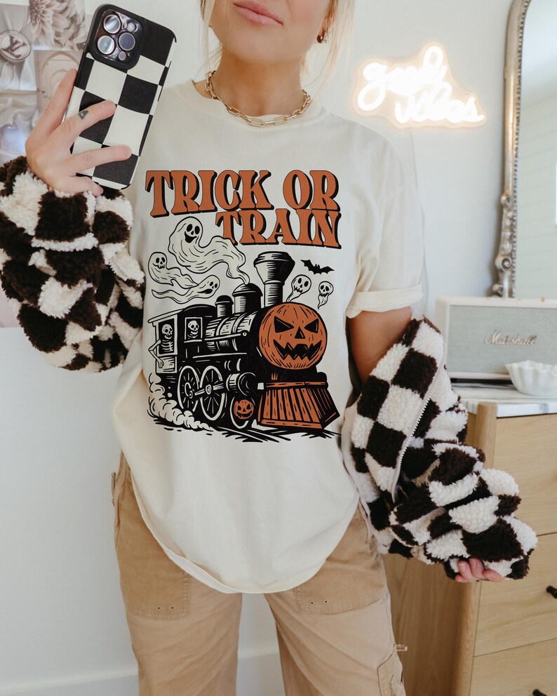 May include: A cream-coloured t-shirt with a Halloween graphic. The design shows a train with a pumpkin face, ghosts, and the text "TRICK OR TRAIN" in orange. The person is wearing a black and white chequered jacket and holding a chequered phone case.