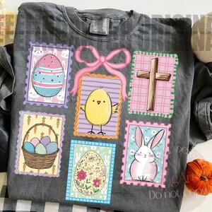 May include: Dark gray t-shirt featuring Easter-themed postage stamp designs. The stamps include a decorated egg, a yellow chick, a cross, a basket of eggs, a floral egg, and a pink bunny. The shirt has a Comfort Colors tag.