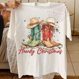May include: White t-shirt featuring a graphic of two cowboy boots, one green and blue, the other red and tan, adorned with Christmas lights and cowboy hats. The text "Howdy Christmas" is written in red script.