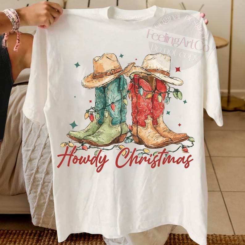 May include: White t-shirt featuring a graphic of two cowboy boots, one green and blue, the other red and tan, adorned with Christmas lights and cowboy hats. The text "Howdy Christmas" is written in red script.