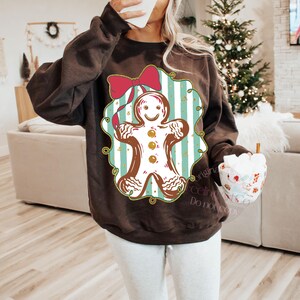 May include: A brown sweatshirt featuring a gingerbread man design. The gingerbread man is white with brown details and gold buttons, set against a blue and white striped background. A red bow is at the top of the design.