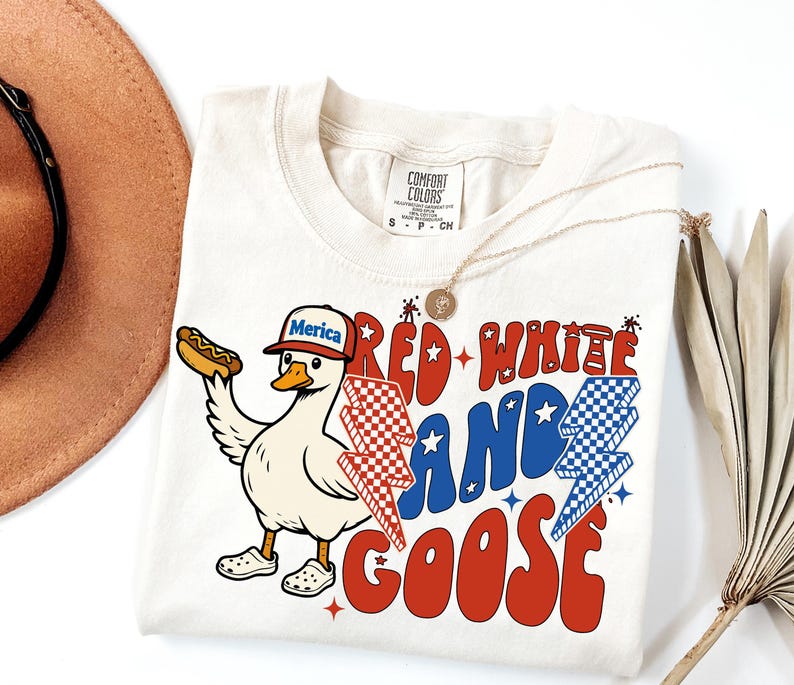 May include: Cream-coloured t-shirt with a graphic of a goose wearing a hat that says "Merica" and holding a hot dog. The shirt features the text "Red, White, and Goose" in red and blue, with lightning bolt designs. A gold necklace is draped on the shirt.
