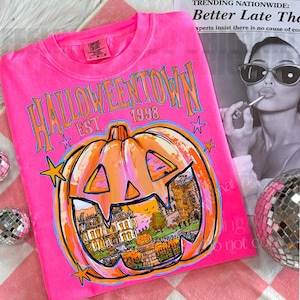 May include: A neon pink t-shirt featuring the text HALLOWEENTOWN EST. 1998 in orange and blue. A large orange jack-o'-lantern graphic contains a detailed illustration of a house and a smaller pumpkin. Small orange stars surround the design.