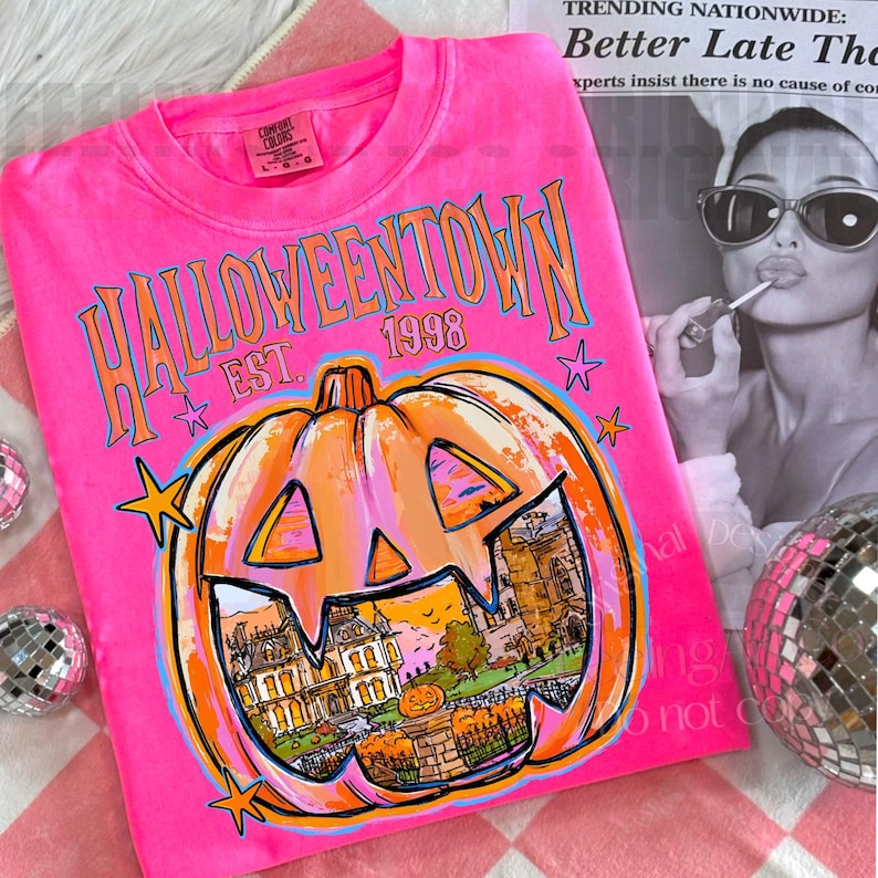 May include: A neon pink t-shirt featuring the text HALLOWEENTOWN EST. 1998 in orange and blue. A large orange jack-o'-lantern graphic contains a detailed illustration of a house and a smaller pumpkin. Small orange stars surround the design.
