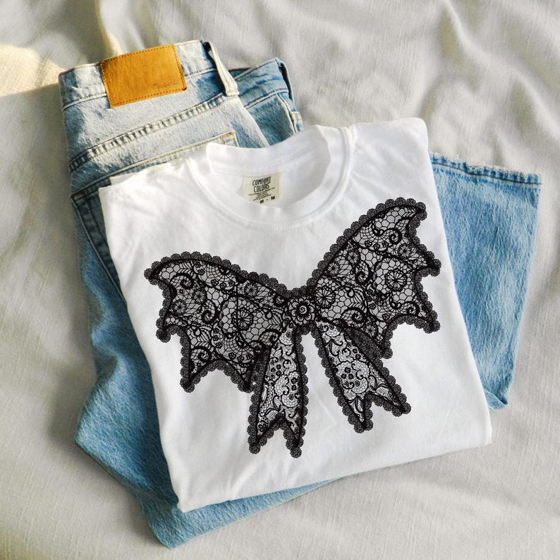 May include: White t-shirt featuring a large black lace bow design. The bow has intricate floral patterns and a scalloped edge. The shirt is folded on top of a pair of light blue denim jeans.