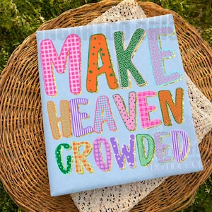 K&ouml;nnte beinhalten: Hellblaues T-Shirt mit dem Schriftzug "MAKE HEAVEN CROWDED" in bunten, gemusterten Buchstaben mit goldenen Glitzerkonturen. Das T-Shirt liegt auf einem Weidenkorb.