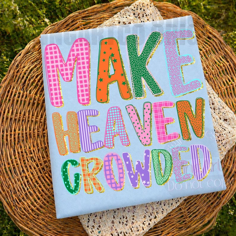 K&ouml;nnte beinhalten: Hellblaues T-Shirt mit dem Schriftzug "MAKE HEAVEN CROWDED" in bunten, gemusterten Buchstaben mit goldenen Glitzerkonturen. Das T-Shirt liegt auf einem Weidenkorb.