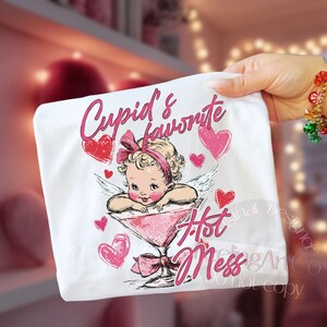 May include: White t-shirt featuring a cartoon cupid in a martini glass, surrounded by pink hearts. The text reads "Cupid's favorite Hot Mess" in pink script. A fun, Valentine's Day themed design.