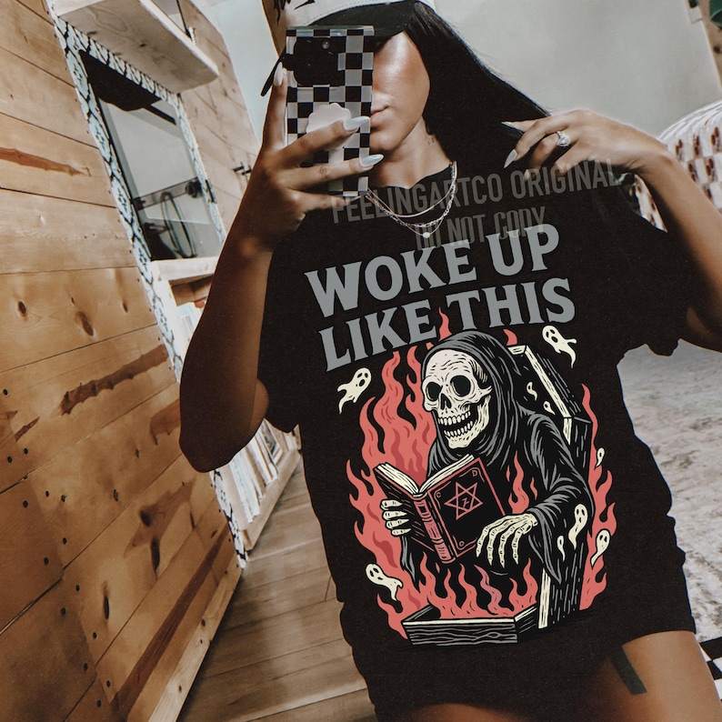 May include: Black t-shirt featuring the phrase "WOKE UP LIKE THIS" and a graphic of a skeleton in a black robe reading a book with a pentagram. The design includes flames and ghost illustrations.