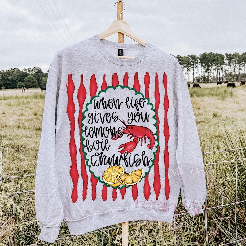 K&ouml;nnte beinhalten: Ein meliertes graues Sweatshirt mit einem Design mit dem Text "Wenn das Leben dir Zitronen gibt, koche Krebse". Das Design umfasst einen roten Krebs, Zitronenscheiben und rote vertikale Streifen.