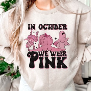 May include: A light beige sweatshirt with a graphic print of a mummy, a ghost, and a pumpkin. The text "In October We Wear Pink" is printed in black letters.