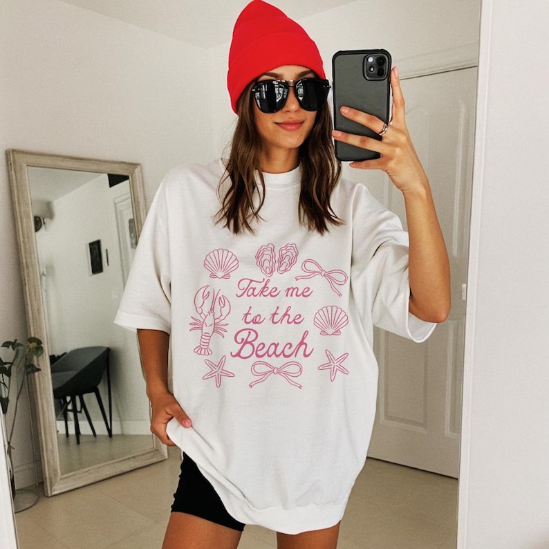 May include: White t-shirt with a pink beach-themed graphic, including seashells, a lobster, and the text "Take me to the Beach." The person is wearing a red beanie and sunglasses.