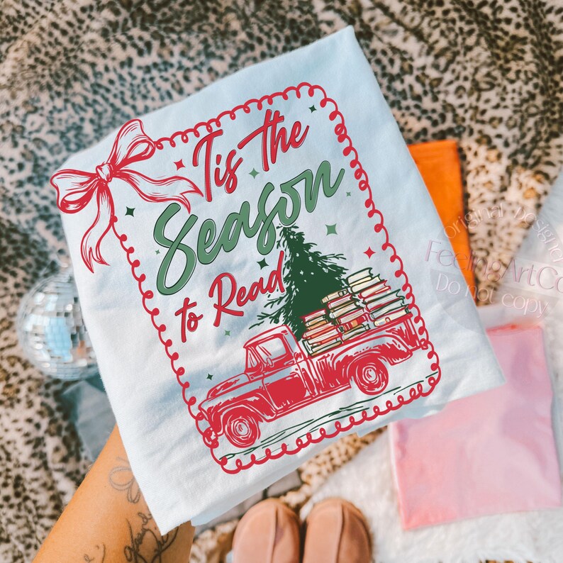 May include: A light blue t-shirt featuring a red truck laden with books and a Christmas tree. The shirt displays the text "Tis the Season to Read" in green and red. A red bow adorns the top left corner of the shirt.