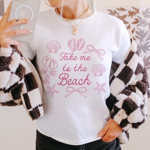 May include: White t-shirt with a pink graphic that says "Take me to the Beach." The graphic includes illustrations of seashells, a lobster, starfish, and bows. The person is wearing a black and white checkered jacket.