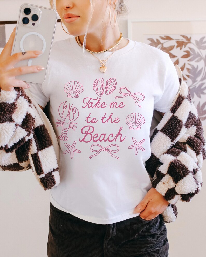 May include: White t-shirt with a pink graphic that says "Take me to the Beach." The graphic includes illustrations of seashells, a lobster, starfish, and bows. The person is wearing a black and white checkered jacket.