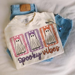 May include: Cream-colored t-shirt with a graphic of three cartoon ghosts wearing bows, each in a different color frame. The text "Spooky Vibes" is below the ghosts. The shirt is paired with light blue jeans and glasses.