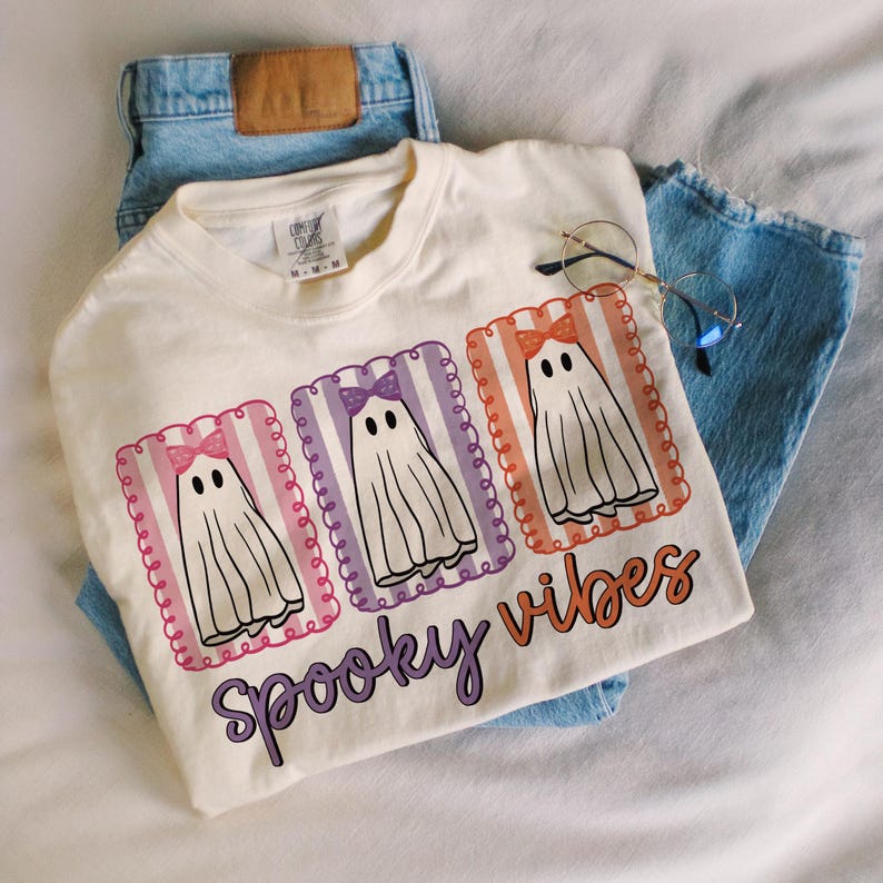 May include: Cream-colored t-shirt with a graphic of three cartoon ghosts wearing bows, each in a different color frame. The text "Spooky Vibes" is below the ghosts. The shirt is paired with light blue jeans and glasses.
