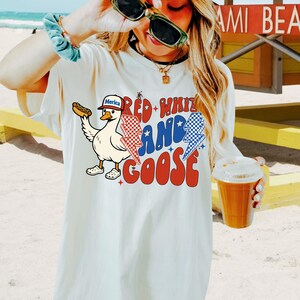 May include: Off-white t-shirt with a cartoon goose holding a hot dog, wearing a hat that says "Merica". The shirt has the text "Red, White, and Goose" in red and blue, with lightning bolt graphics. The design is patriotic.
