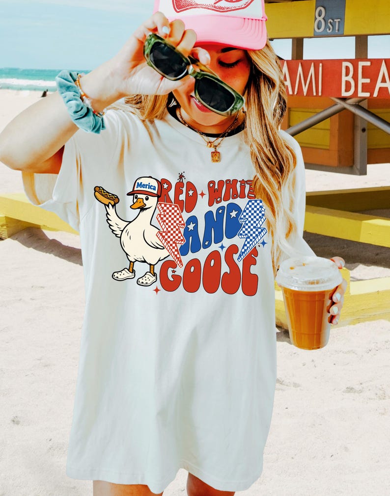 May include: Off-white t-shirt with a cartoon goose holding a hot dog, wearing a hat that says "Merica". The shirt has the text "Red, White, and Goose" in red and blue, with lightning bolt graphics. The design is patriotic.