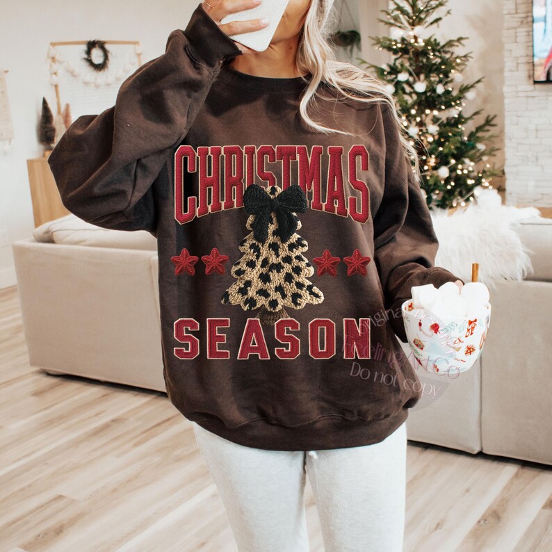May include: A brown sweatshirt with the words "CHRISTMAS SEASON" in red block letters. A leopard print Christmas tree with a black bow is centered on the sweatshirt. Red stars are on either side of the tree.