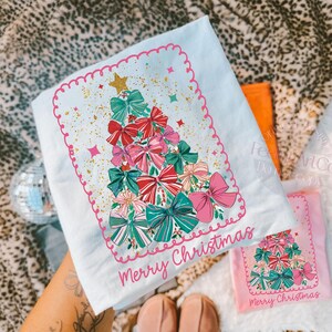 May include: A light blue Christmas t-shirt with a festive design. The shirt features a Christmas tree composed of colourful bows in shades of red, pink, and green, with gold glitter and the words "Merry Christmas".