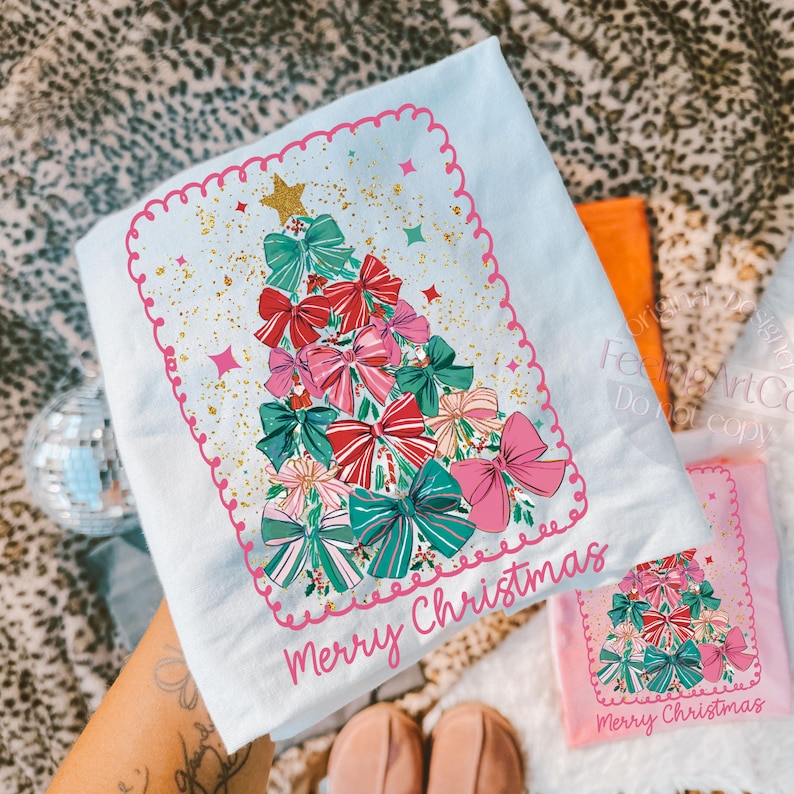 May include: A light blue Christmas t-shirt with a festive design. The shirt features a Christmas tree composed of colourful bows in shades of red, pink, and green, with gold glitter and the words "Merry Christmas".