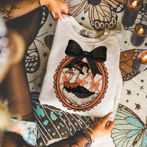 Peut inclure: Sweat-shirt cr&egrave;me avec une illustration fantaisiste d'une oie portant un chapeau de sorci&egrave;re et un col &agrave; volants, dans un cadre festonn&eacute; orange. Un grand n&oelig;ud noir orne le haut. Th&egrave;me d'Halloween.
