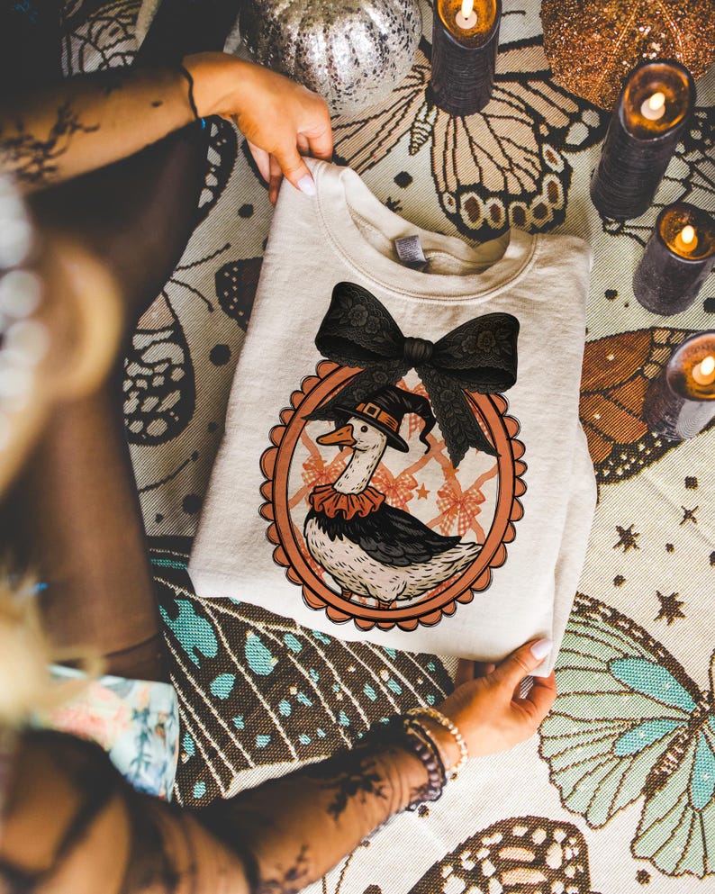 Peut inclure: Sweat-shirt cr&egrave;me avec une illustration fantaisiste d'une oie portant un chapeau de sorci&egrave;re et un col &agrave; volants, dans un cadre festonn&eacute; orange. Un grand n&oelig;ud noir orne le haut. Th&egrave;me d'Halloween.