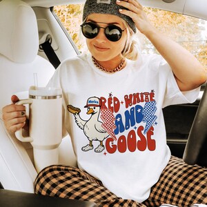 May include: White t-shirt with a graphic of a goose wearing a hat and holding a hot dog, with the text "Red, White, and Goose." The shirt is paired with plaid trousers, a grey beanie, and sunglasses. A large white mug is also visible.