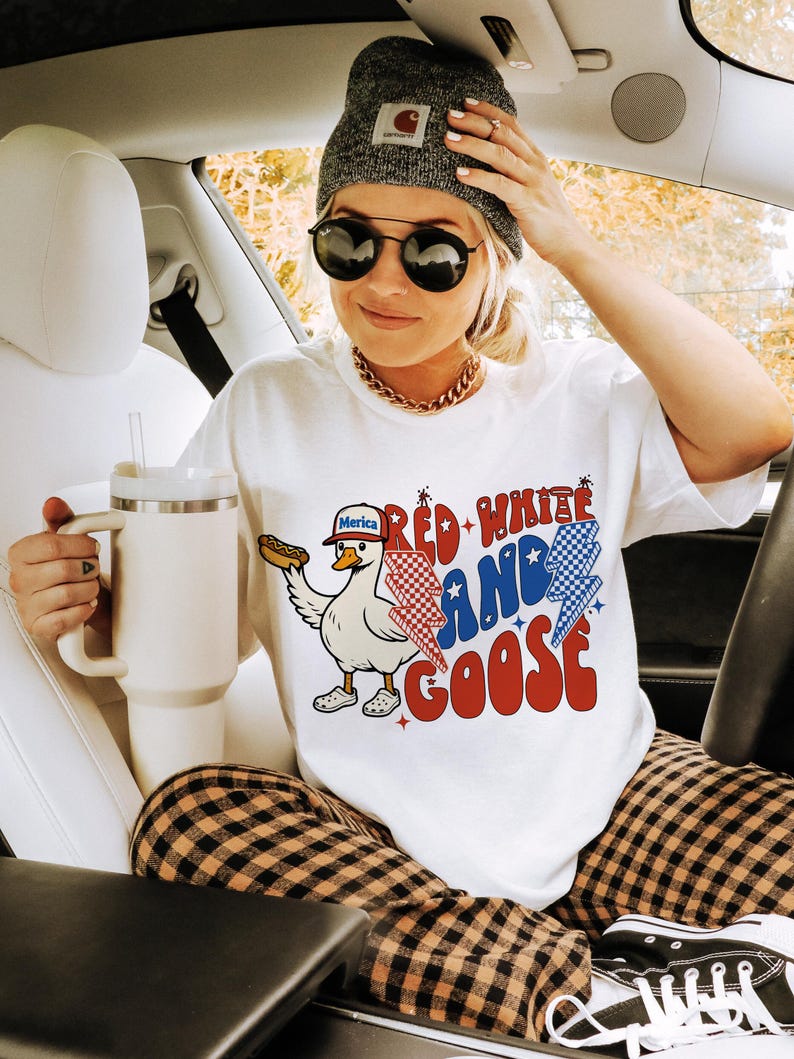 May include: White t-shirt with a graphic of a goose wearing a hat and holding a hot dog, with the text "Red, White, and Goose." The shirt is paired with plaid trousers, a grey beanie, and sunglasses. A large white mug is also visible.