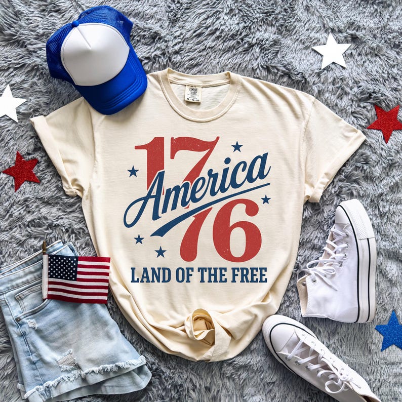 May include: Cream-coloured t-shirt with the text "17 America 76 Land of the Free" in red and blue. The image includes a blue and white trucker hat, denim shorts, a small American flag, and white trainers.