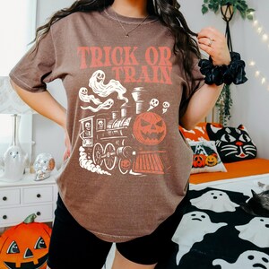 May include: A brown t-shirt with the words "TRICK OR TRAIN" in orange. The graphic depicts a Halloween train with a pumpkin, ghosts, and a skull. The shirt is a relaxed fit, ideal for the spooky season and Halloween.