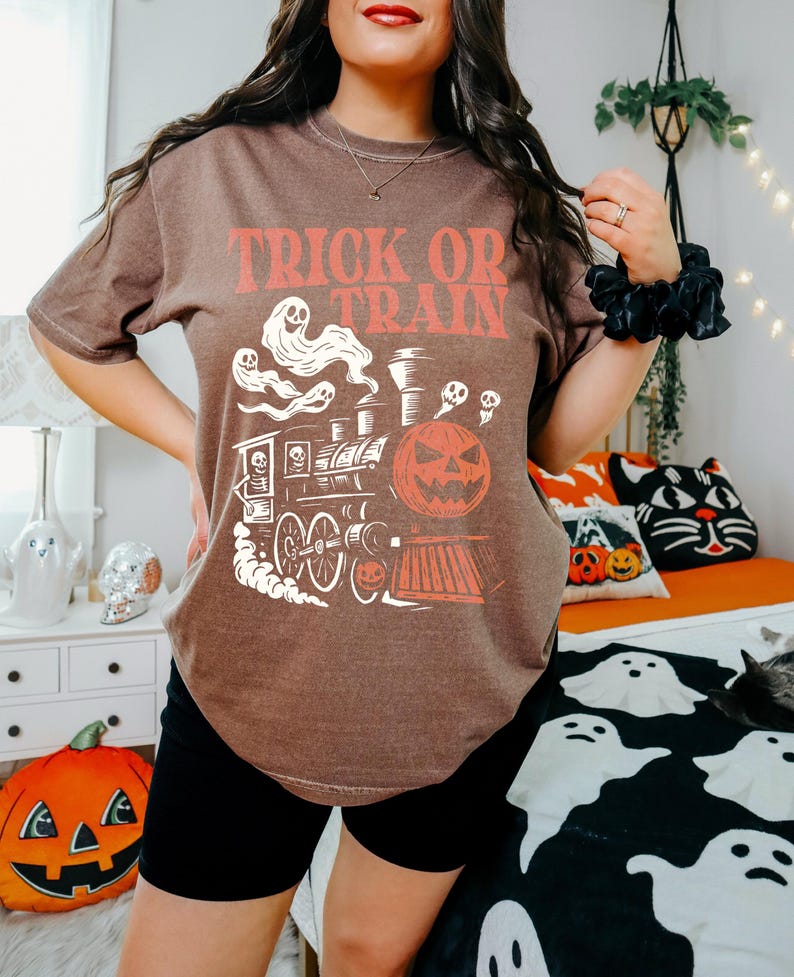 May include: A brown t-shirt with the words "TRICK OR TRAIN" in orange. The graphic depicts a Halloween train with a pumpkin, ghosts, and a skull. The shirt is a relaxed fit, ideal for the spooky season and Halloween.