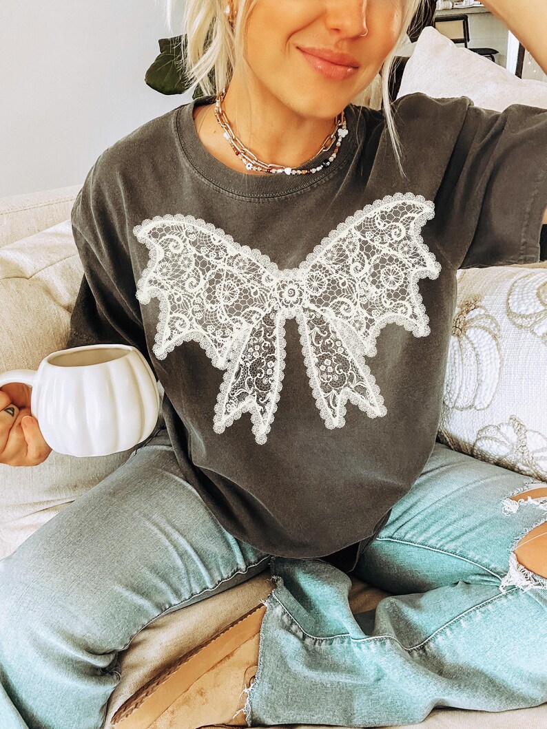 May include: Dark gray t-shirt featuring a large white lace bow design. The shirt has short sleeves and a crew neck. The model is wearing light blue jeans and holding a white pumpkin-shaped mug. The shirt is a casual, comfortable style.