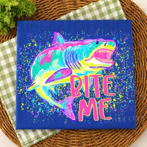 Op de afbeelding: Een blauw canvas toont een kleurrijke haaienillustratie met de tekst "BITE ME" in roze. De haai is afgebeeld in tinten roze, geel, groen en blauw, tegen een achtergrond van kleurrijke verfspetters. Het canvas ligt op een geweven mat.