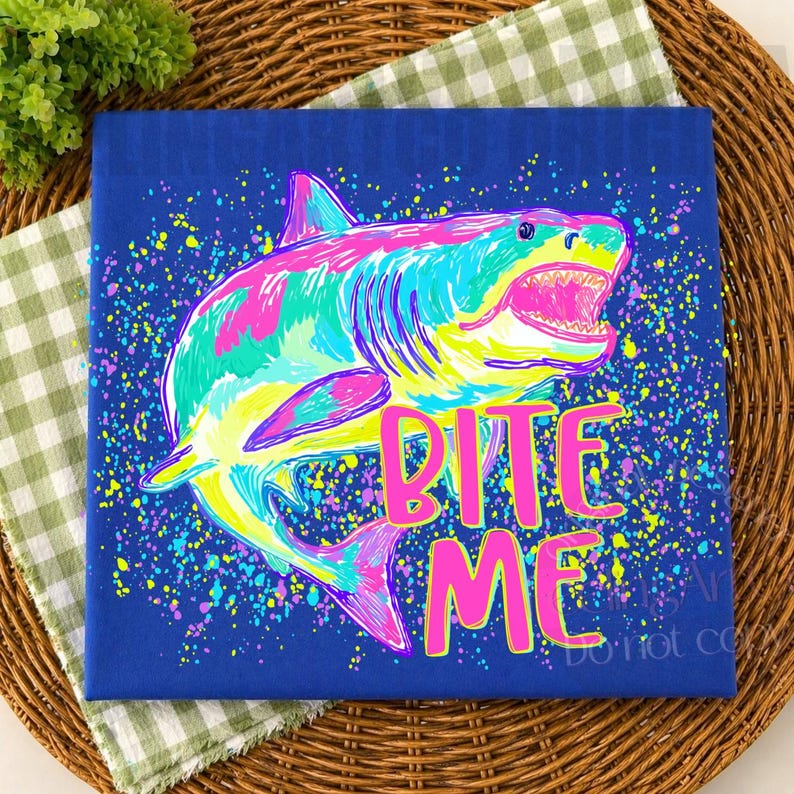 Op de afbeelding: Een blauw canvas toont een kleurrijke haaienillustratie met de tekst "BITE ME" in roze. De haai is afgebeeld in tinten roze, geel, groen en blauw, tegen een achtergrond van kleurrijke verfspetters. Het canvas ligt op een geweven mat.