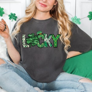 May include: Dark gray t-shirt with the word "LUCKY" in a green and white design. The letter "L" has a leopard print pattern, the "U" is a shamrock, and the "Y" has a horseshoe design. The shirt is a casual style.