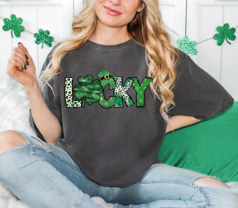 May include: Dark gray t-shirt with the word "LUCKY" in a green and white design. The letter "L" has a leopard print pattern, the "U" is a shamrock, and the "Y" has a horseshoe design. The shirt is a casual style.