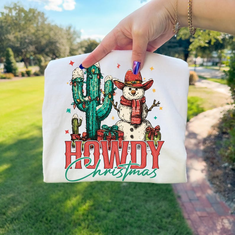 Peut inclure: Sweat-shirt blanc avec un motif color&eacute; repr&eacute;sentant un cactus d&eacute;cor&eacute; de lumi&egrave;res de No&euml;l et un bonhomme de neige portant un chapeau de cowboy et une &eacute;charpe. Le texte "HOWDY Christmas" est affich&eacute; en rouge et vert.