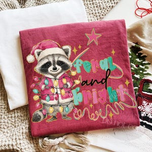 May include: A maroon t-shirt featuring a raccoon wearing a Santa hat and a jacket adorned with Christmas lights. The text "feral and bright" is written in teal, pink, and black. The design includes a star and a swirl of lights.