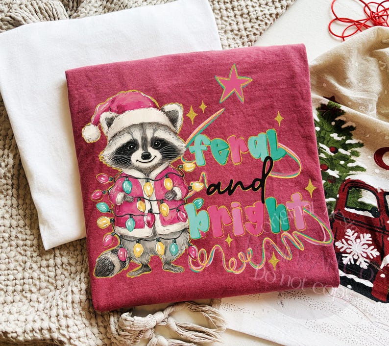 May include: A maroon t-shirt featuring a raccoon wearing a Santa hat and a jacket adorned with Christmas lights. The text "feral and bright" is written in teal, pink, and black. The design includes a star and a swirl of lights.