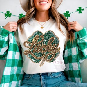 May include: White crew neck t-shirt with a large, sparkling green and gold shamrock design. The word "Lucky" is written across the shamrock in gold script. The person is wearing a green and white plaid shirt and blue jeans.